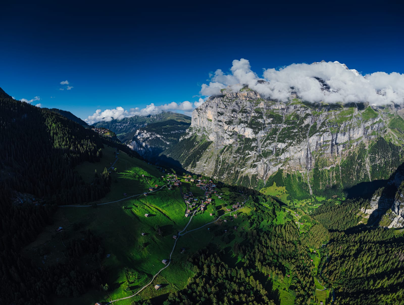 Aerial: alpine valley, limestone cliff wall, village below.